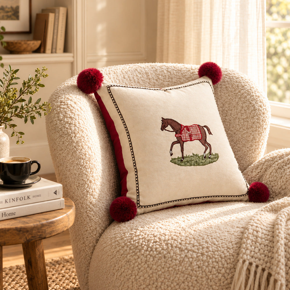 Angled view of cream horse pillow cover with red pom poms on a textured armchair
