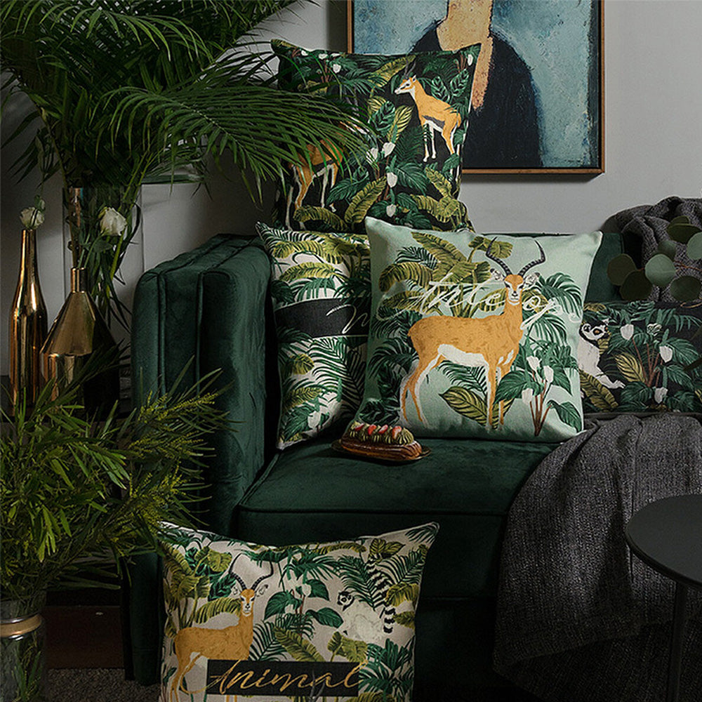 Two tropical jungle pillow covers styled on a dark green velvet sofa with lush indoor plants.