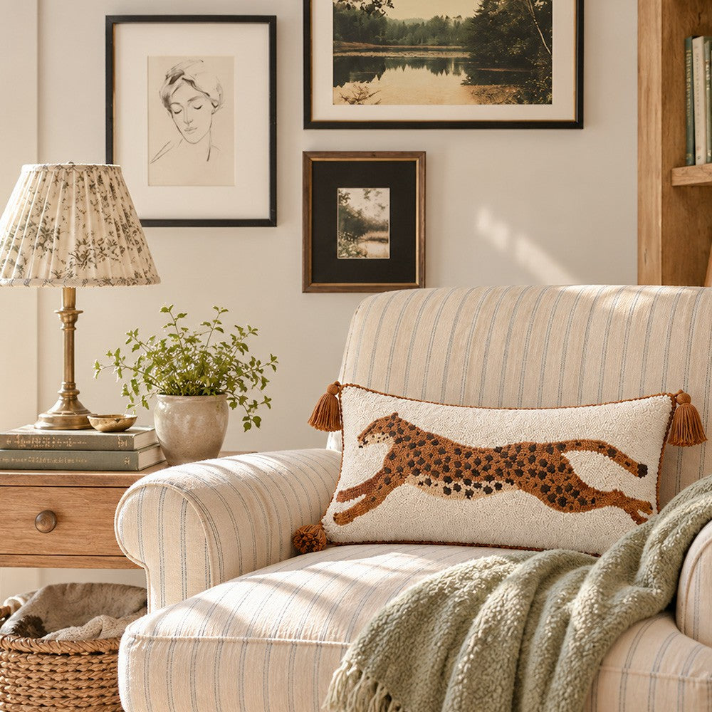 A cream and beige striped armchair in a styled living room, featuring a decorative throw pillow with a brown and black leopard print. A patterned table lamp, a small potted plant, and a woven basket sit on the side table next to the chair, with framed wall art in the background.