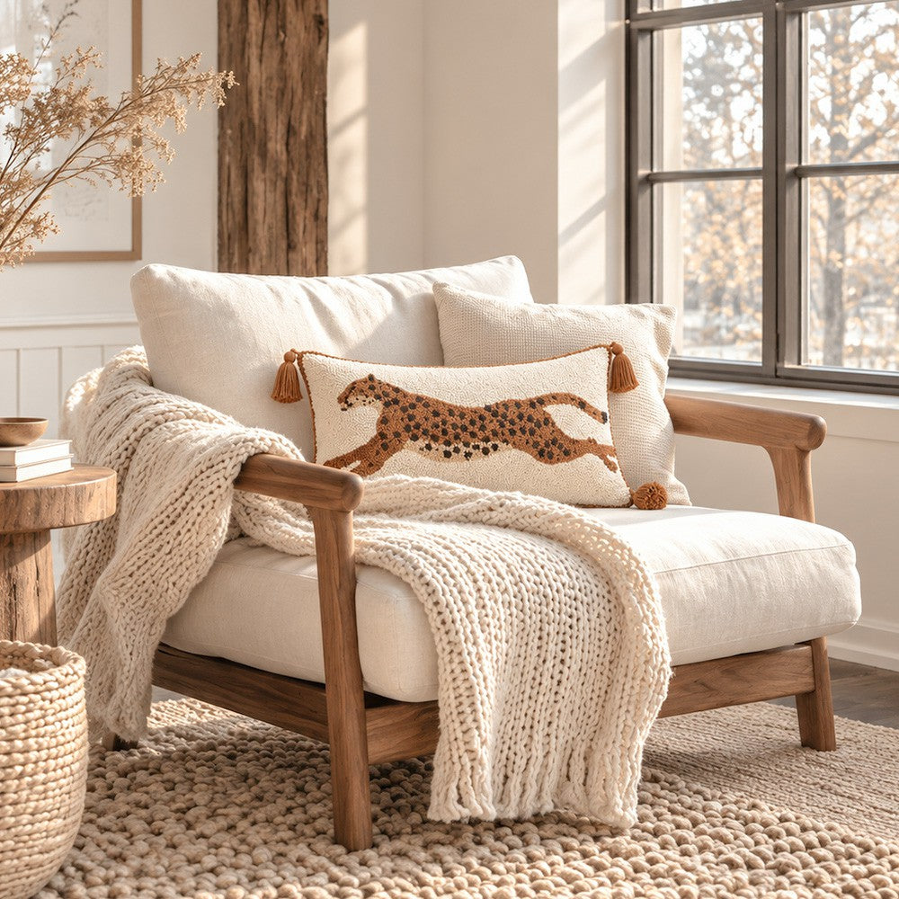 A cozy cream upholstered armchair with a wooden frame, styled with a chunky knit cream throw blanket and a decorative throw pillow featuring a brown spotted animal motif, set in a bright, sunlit room with a window and rustic decor.