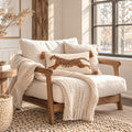 A cozy cream upholstered armchair with a wooden frame, styled with a chunky knit cream throw blanket and a decorative throw pillow featuring a brown spotted animal motif, set in a bright, sunlit room with a window and rustic decor.